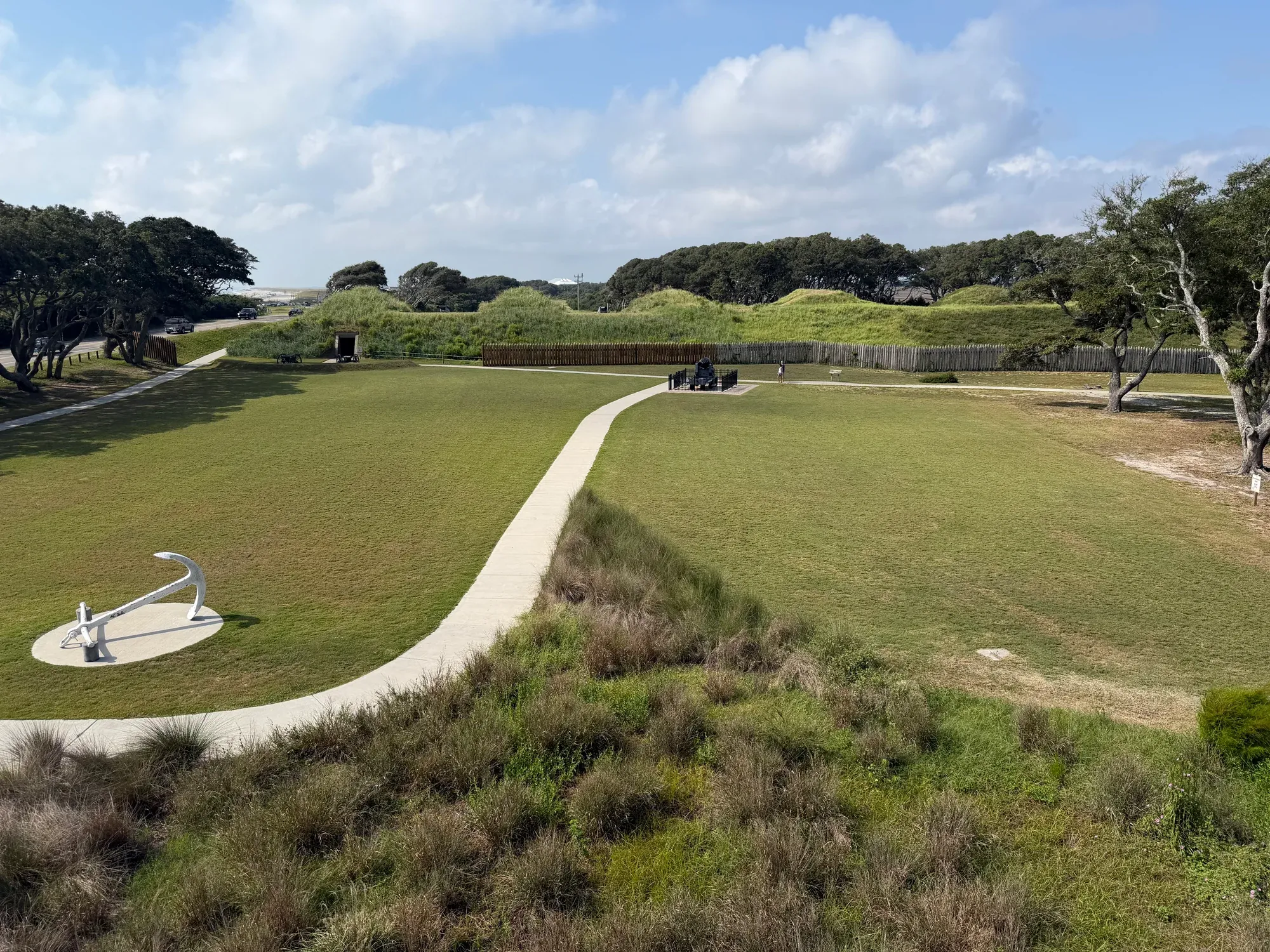 A broad view of Fort Fisher's grass-covered earthworks, wooden palisade, lawn, and curved paved path.